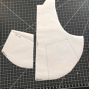 May include: Two white paper patterns for sewing a garment. The larger pattern has a curved shape and the smaller pattern is a curved triangle. The patterns are on a black cutting mat with white grid lines. The text "Fold Line" and "<&larr; Fold" are written on the patterns. The text "OMKCA 2021" is written on the bottom right corner of the mat.
