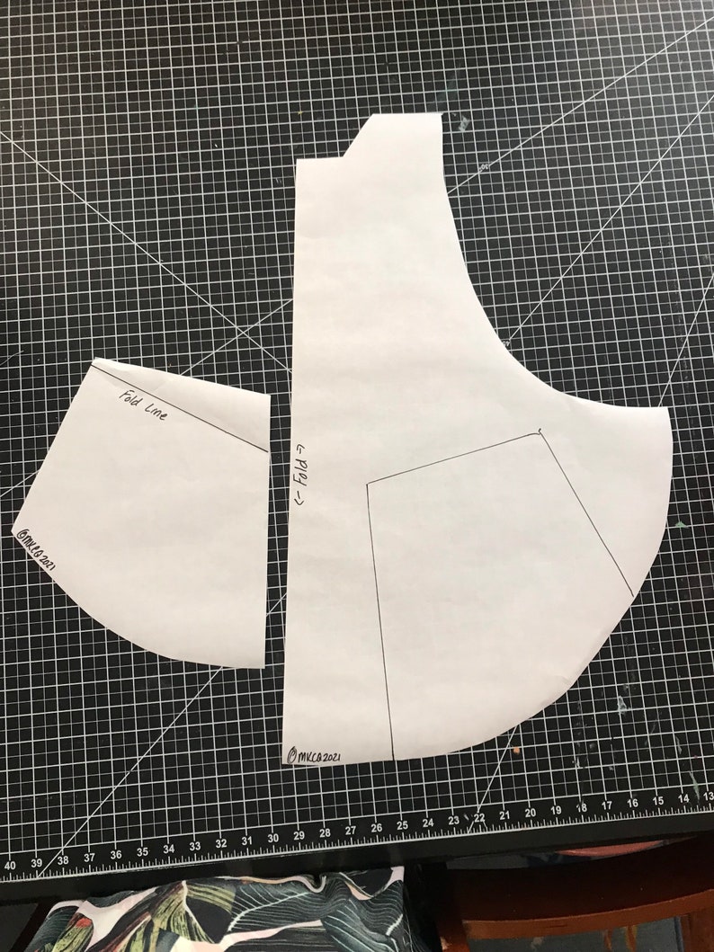 May include: Two white paper patterns for sewing a garment. The larger pattern has a curved shape and the smaller pattern is a curved triangle. The patterns are on a black cutting mat with white grid lines. The text "Fold Line" and "<&larr; Fold" are written on the patterns. The text "OMKCA 2021" is written on the bottom right corner of the mat.