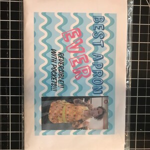 May include: A white and blue patterned apron with the text "Best Apron Ever" and "Reversible!!! With Pockets!!!". The apron is being worn by a person with tattoos.