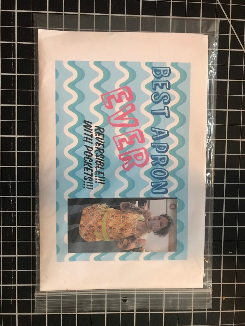 May include: A white and blue patterned apron with the text "Best Apron Ever" and "Reversible!!! With Pockets!!!". The apron is being worn by a person with tattoos.