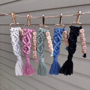 May include: Six macrame keychains in various colors, including gray, pink, green, blue, and black. Each keychain features a gold-colored clasp, wooden beads spelling out names, and a matching tassel. The keychains are hanging on a string.