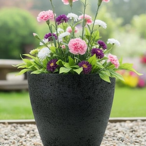 May include: A black, textured plant pot overflowing with a vibrant mix of flowers. The arrangement features pink carnations, purple, white, and yellow blooms, and green leaves. The pot rests on a bed of small pebbles.