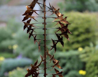 Op de afbeelding: Een metalen tuinsculptuur van een zwerm vogels in de vlucht. De vogels zijn gemaakt van koper en zijn in een spiraalvorm gerangschikt. De sculptuur is gemonteerd op een metalen paal.