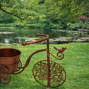 May include: A copper-finished metal bicycle planter. The front wheel and the sides of the planter feature a tree design. A small bird adorns the handlebars. The planter is set on a grassy lawn, with a pond in the background.