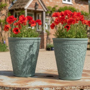 May include: Two sage green flower pots, each filled with red poppies. The pots have an embossed floral design and sit on a weathered metal table. The background features a brick building and a wooden fence.