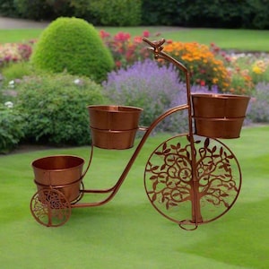 May include: A bronze metal bicycle planter with three bucket-shaped pots. The bicycle features a decorative tree of life design on the wheel. The planter is set on a green lawn, with a garden and flowers in the background.