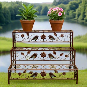 May include: A three-tiered, brown metal plant stand with decorative bird and floral accents. The stand holds two terracotta pots, one with green foliage and the other with pink flowers. The stand is outdoors near a lake.
