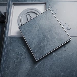Puede incluir: Una rejilla de ducha cuadrada con textura de piedra gris oscuro y marco metálico. La rejilla está parcialmente levantada, revelando un componente de drenaje circular gris claro debajo. Se observan varias gotas de agua en la superficie.
