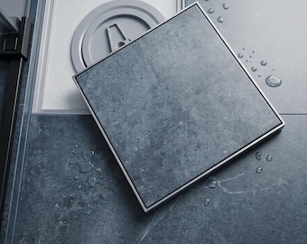Può includere: Una copertura quadrata per scarico doccia con texture effetto pietra grigio scuro e cornice metallica. La copertura è parzialmente sollevata, rivelando un componente di scarico circolare grigio chiaro, su un pavimento piastrellato grigio scuro con gocce d'acqua.