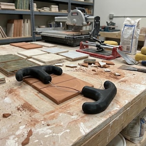 Puede incluir: Una escena de taller con varias baldosas de diferentes colores y tamaños. Una sierra para baldosas, lechada y herramientas están sobre una mesa de madera. Dos cortadores de baldosas negros están sobre una baldosa de terracota.