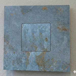 Puede incluir: Una baldosa cuadrada de piedra color pizarra con un área rectangular empotrada en el centro. La baldosa tiene un aspecto moteado con tonos de azul, gris y toques de dorado. La piedra tiene una superficie natural y texturizada.