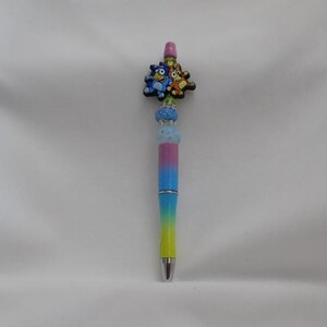 May include: A decorative pen with a colorful design. The pen features a gradient of colors, transitioning from yellow to blue, with a silver tip. The top of the pen is adorned with a charm featuring cartoon characters.