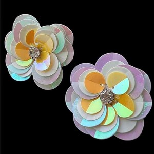 Peut inclure: Deux accessoires en forme de fleur avec des paillettes irisées. Chaque fleur a une gemme centrale et des pétales dans les tons de blanc, jaune, orange et bleu. Le fond est noir.