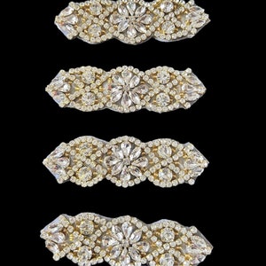 Può includere: Quattro applicazioni decorative con strass. Ogni applicazione presenta un motivo floreale centrale circondato da strass più piccoli e scintillanti. Le applicazioni hanno un bordo dorato e sono montate su un supporto in tessuto colorato chiaro.