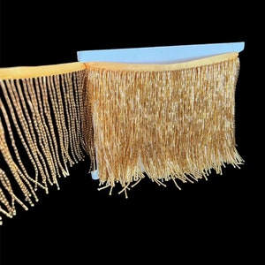 Peut inclure: Garniture de franges dorées avec une texture perlée dense. La garniture est fixée à un support de couleur claire et pend sur un fond noir. La frange est composée de nombreux brins de perles dorées.