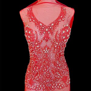 Puede incluir: Una prenda roja sin mangas con intrincados abalorios y adornos de pedrería. El diseño presenta un cuello en V y un patrón floral detallado en el corpiño. La tela transparente está drapeada sobre un maniquí.