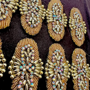 May include: Close-up of several gold-colored, oval-shaped embellishments. Each features a beaded border, a central cluster of iridescent rhinestones, and metallic accents. The embellishments are arranged on a dark background.