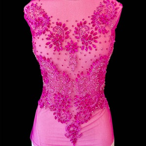 Puede incluir: Una blusa rosa sin mangas con intrincados abalorios y adornos. La blusa presenta un cuello alto y está adornada con patrones florales y de estilo paisley en tonos de rosa y magenta. El diseño está centrado en la parte delantera de la prenda.