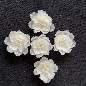 Pode incluir: Cinco enfeites de flores de tecido branco translúcido com centros perolados. As flores estão dispostas sobre um fundo escuro e texturizado. Cada flor tem múltiplas pétalas e um grupo de pequenas contas redondas no centro.