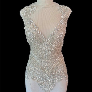 May include: A sleeveless, cream-colored garment with intricate beadwork. The bodice features a deep V-neck and is heavily embellished with clear, sparkling beads and rhinestones in floral and geometric patterns. The garment is displayed on a mannequin against a black background.