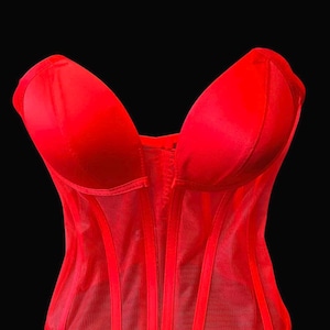 May include: A vibrant red corset with a structured bodice and a sheer mesh overlay. The corset features a sweetheart neckline and vertical boning for a defined silhouette. The garment is set against a black background.