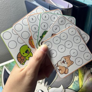 May include: A set of square paper savings trackers featuring cartoon animals including a turtle, lion, frog, and dog. Each card has a grid of circles with printed numbers 1, 2, or 3, and the text eiskabudgets is visible on the bottom corner of the cards.