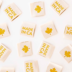 Puede incluir: Un fondo blanco con múltiples etiquetas de tela beige con letras doradas brillantes. Las etiquetas dicen "Made in CA" y tienen una hoja de arce dorada brillante.