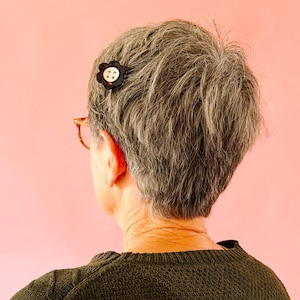 May include: A black flower-shaped hair clip with a white button center is pinned in gray hair. The person is wearing a dark green sweater and glasses. The background is a solid pink.