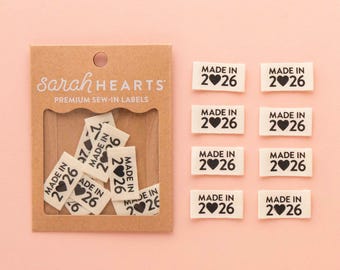 May include: A collection of white sew-in labels with black text that reads "MADE IN 2❤26". The labels are displayed on a light pink background, with some labels inside a brown cardboard package that says "Sarah HEARTS PREMIUM SEW-IN LABELS".