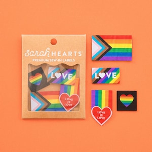 Puede incluir: Un conjunto de etiquetas para coser premium Sarah Hearts en un paquete de cartón marrón con ventana. Las etiquetas presentan diseños de bandera arcoíris, formas de corazón y el texto "LOVE" o "LOVE IS LOVE" en blanco sobre fondos coloridos.