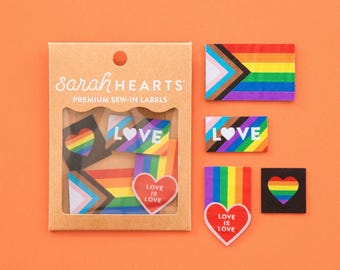 May include: A set of Sarah Hearts premium sew-in labels in a brown cardboard package with a window. The labels feature rainbow pride flag designs, heart shapes, and the text "LOVE" or "LOVE IS LOVE" in white on various colorful backgrounds.