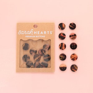 May include: A package of "Sarah Hearts Premium Buttons" with a clear window, displaying several round, tortoiseshell-patterned buttons. Additional buttons are scattered on a pink surface. The buttons have a mix of brown, black, and amber tones.