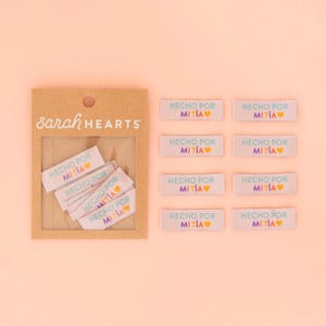May include: A pack of 10 white woven labels with the text "HECHO POR MITIA" in rainbow colors and a heart. The labels are in a brown cardboard box with the text "Sarah HEARTS" on the front.