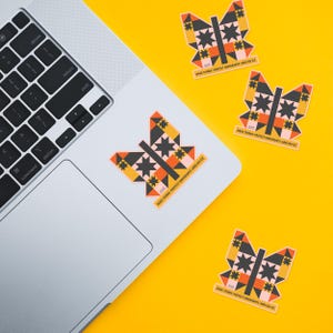 May include: Butterfly-shaped stickers with geometric designs in black, orange, yellow, and pink are scattered on a yellow surface and a laptop. The stickers have the text "MAKE THINGS - PROTECT IMMIGRANTS - ABOLISH ICE".
