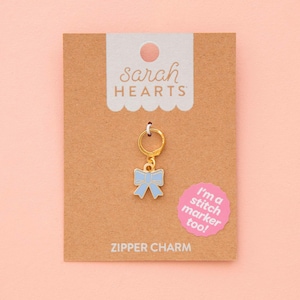 May include: A gold-colored zipper charm with a light blue bow pendant. The charm is attached to a brown card with the text "ZIPPER CHARM" and the brand name "sarah HEARTS". A pink sticker says "I'm a stitch marker too!"