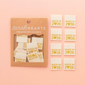 May include: A collection of cream-colored sew-in labels with gold text and a heart design. The labels read "MADE IN 2026". The labels are packaged in a brown cardboard box with the text "Sarah HEARTS PREMIUM SEW-IN LABELS".