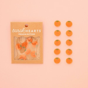 May include: A set of orange tomato-shaped buttons with green stems, packaged in a brown cardboard card labeled sarah HEARTS PREMIUM BUTTONS, alongside ten loose matching buttons arranged in two vertical columns of five on a light surface.