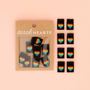 Peut inclure: Un paquet de 10 étiquettes en tissu noir avec des cœurs arc-en-ciel. Les étiquettes sont emballées dans une boîte en carton marron avec le texte "Sarah Hearts" imprimé sur le devant.