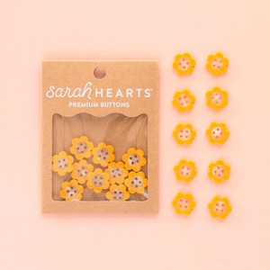 May include: A collection of yellow flower-shaped buttons with brown centers. The buttons are displayed both in a clear plastic package and scattered on a light pink surface. The package reads "sarah HEARTS PREMIUM BUTTONS."