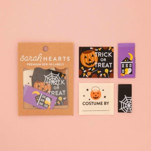 Op de afbeelding: Een set Halloween-labels om in te naaien van Sarah Hearts in een bruine kartonnen verpakking. De labels bevatten ontwerpen van pompoenen, spinnenwebben, een spookhuis en teksten zoals Trick or Treat en Costume By in zwart, oranje en paars.