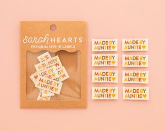 Op de afbeelding: Een bruine kartonnen verpakkingskaart met het label sarah HEARTS PREMIUM SEW-IN LABELS met daarin diverse kleine rechthoekige stoffen labels bedrukt met de tekst MADE BY AUNTIE in oranje, rode en groene letters, inclusief acht losse bijpassende labels.