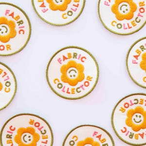 Peut inclure: Écussons circulaires brodés avec le texte "FABRIC COLLECTOR" en rose et orange. Chaque écusson présente un motif de fleur souriante au centre. Les écussons ont un fond blanc et une bordure verte.