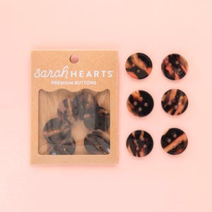 May include: A package of brown and black buttons with a tortoiseshell pattern, next to a few loose buttons. The package reads "sarah HEARTS PREMIUM BUTTONS" on a light brown background. The buttons are round with four holes.