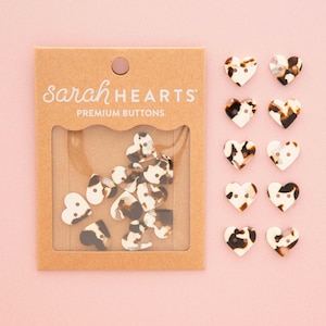 May include: A set of heart-shaped buttons with a brown, black, and white tortoiseshell pattern. The buttons are packaged in a brown cardboard card labeled sarah HEARTS PREMIUM BUTTONS with a clear window displaying several loose buttons.