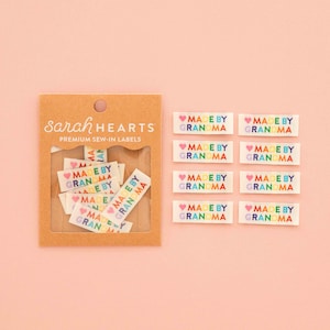 以下が含まれることがあります： Sarah Hearts Premium Sew-In Labelsと書かれた茶色の厚紙パッケージ。ピンクのハートと虹色の文字でMADE BY GRANDMAとプリントされた白い布製ラベルが数枚入っており、横には同じラベルが8枚並べられています。