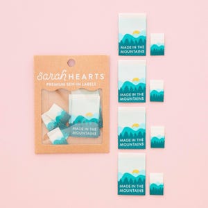 Pode incluir: Uma embalagem de cartão castanho com a etiqueta Sarah Hearts Premium Sew-In Labels contendo várias etiquetas de tecido retangulares. Cada etiqueta apresenta um gráfico de montanhas verdes, um sol amarelo e o texto Made in the Mountains em branco.