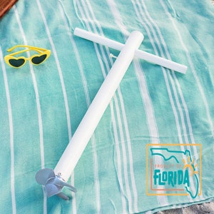 May include: White sand anchor with a silver propeller, lying on a blue and white striped beach towel. The anchor has a white T-shaped handle. The towel has the words "Property of Florida" printed on it.