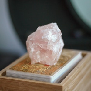 May include: A raw, translucent pale pink rose quartz crystal chunk resting on top of a deck of rectangular playing cards with ornate gold and brown patterned backs, all contained within a light-colored wooden box with visible grain.