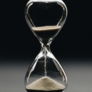 May include: A glass hourglass with sand flowing from the top bulb to the bottom bulb. The hourglass is on a black surface.