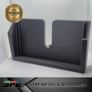 May include: A black Stream Deck XL holder with a rectangular design. The holder has a U-shaped opening and a flat base. The text "STREAM DECK XL HOLDER" is displayed at the bottom. A gold seal with the text "100% PREMIUM QUALITY" is in the upper left corner.
